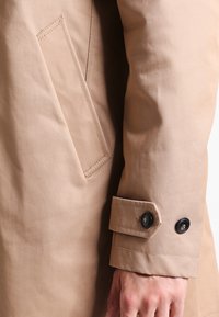 Beige vattentät jacka med sidficka och knäppta ärmslut med svarta knappar. Slät textur och skräddarsydd passform, designad för mångsidighet.