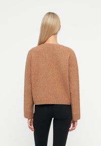 Maglione marrone lavorato a maglia con una vestibilità rilassata, collo rotondo e maniche lunghe, con polsini e orlo a coste. Indossato con pantaloni neri.