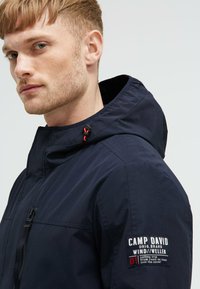 Granatowa wodoodporna kurtka z zamkiem i kapturem, z czerwonymi akcentami, wyszywaną logotypem na rękawie oraz gładką, matową fakturą.
