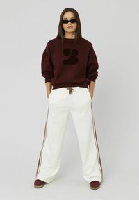 Sweatshirt bordeaux avec un logo sombre, associé à un pantalon large blanc avec des rayures latérales marron et un cordon de serrage. Porté avec des baskets bordeaux.