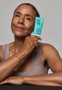 Turquoise tube van NIP+FAB Hyaluronic Fix Nourishing Moisturiser SPF 50 vastgehouden door een persoon, met een matte afwerking en heldere tekstlabel, op een donkere ondergrond.