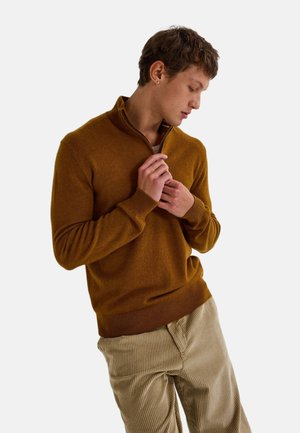 Pull à col zippé marron en matériau doux, présentant une coupe ajustée et des poignets côtelés, associé à un pantalon en velours côtelé beige.