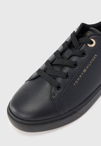 Černé kožené tenisky s černými tkaničkami, zlatými očka a textovým logem "Tommy Hilfiger" v zlaté barvě na boku, umístěné na bílém pozadí.