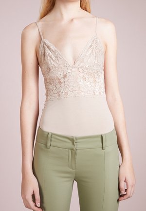 Béžový krajkový top na ramínka s nastavitelnými řemínky, s výstřihem do V, vrstvený přes hladké béžové tílko, doplněný olivově zelenými přiléhavými kalhotami.