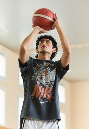 Basketbalspeler in een donker graphic T-shirt met een gevleugeld paard ontwerp, die een rood-witte basketbal schiet, binnen met helder licht.