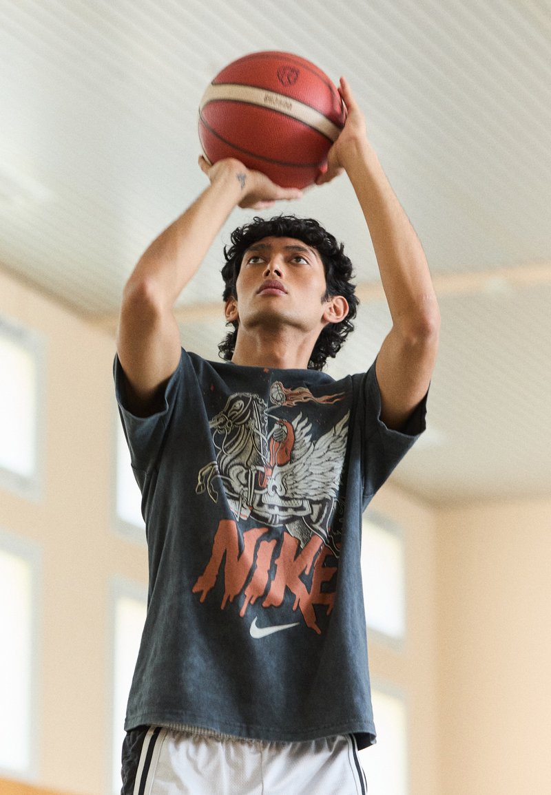 Basketballspieler trägt ein dunkles grafisches T-Shirt mit einem Design eines geflügelten Pferdes, während er einen rot-weißen Basketball wirft, drinnen bei hellem Licht.