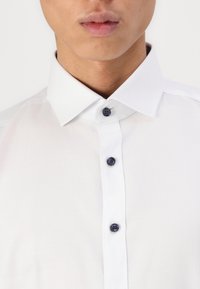Hvit skjorte med knappestolpe som har klassisk krage, blå knapper og teksturert stoff. Nærbilde som fremhever krage- og knappdetaljer.