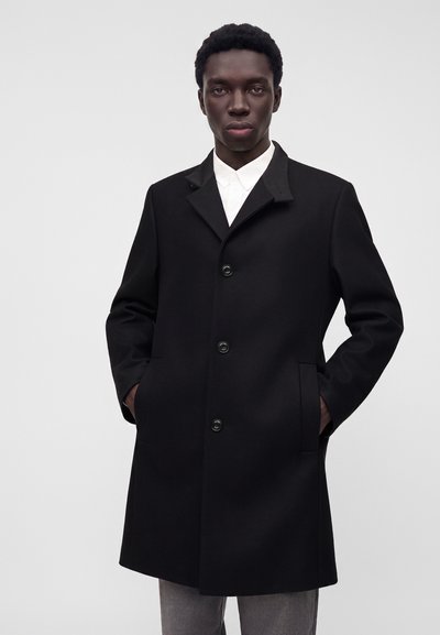 Homme portant un pardessus noir boutonné, les mains dans les poches, chemise blanche et pantalon gris, debout devant un fond uni gris clair.