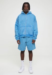 Lyseblå hettegenser med glidelås foran, store lommer og matchende shorts. Laget av mykt materiale, med en avslappet passform og ribbestrikkede ermer.