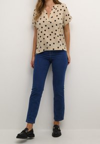 Blouse beige à manches courtes avec des pois noirs, décolleté en V, associée à un jean skinny bleu et des mocassins noirs avec des détails en métal.