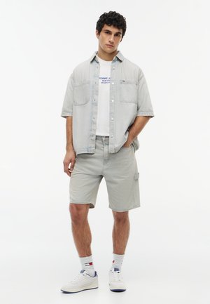 Junger Mann steht, trägt ein hellgraues kurzärmeliges Hemd, passende Shorts, ein weißes bedrucktes T-Shirt, weiße Sneakers und weiße Socken.