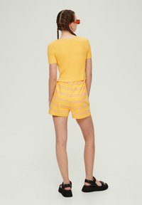 Gelbes ribbeliges T-Shirt mit kurzen Ärmeln, kombiniert mit gelben gestreiften Shorts mit pinken Linien, getragen mit schwarzen Sandalen mit Riemchen.