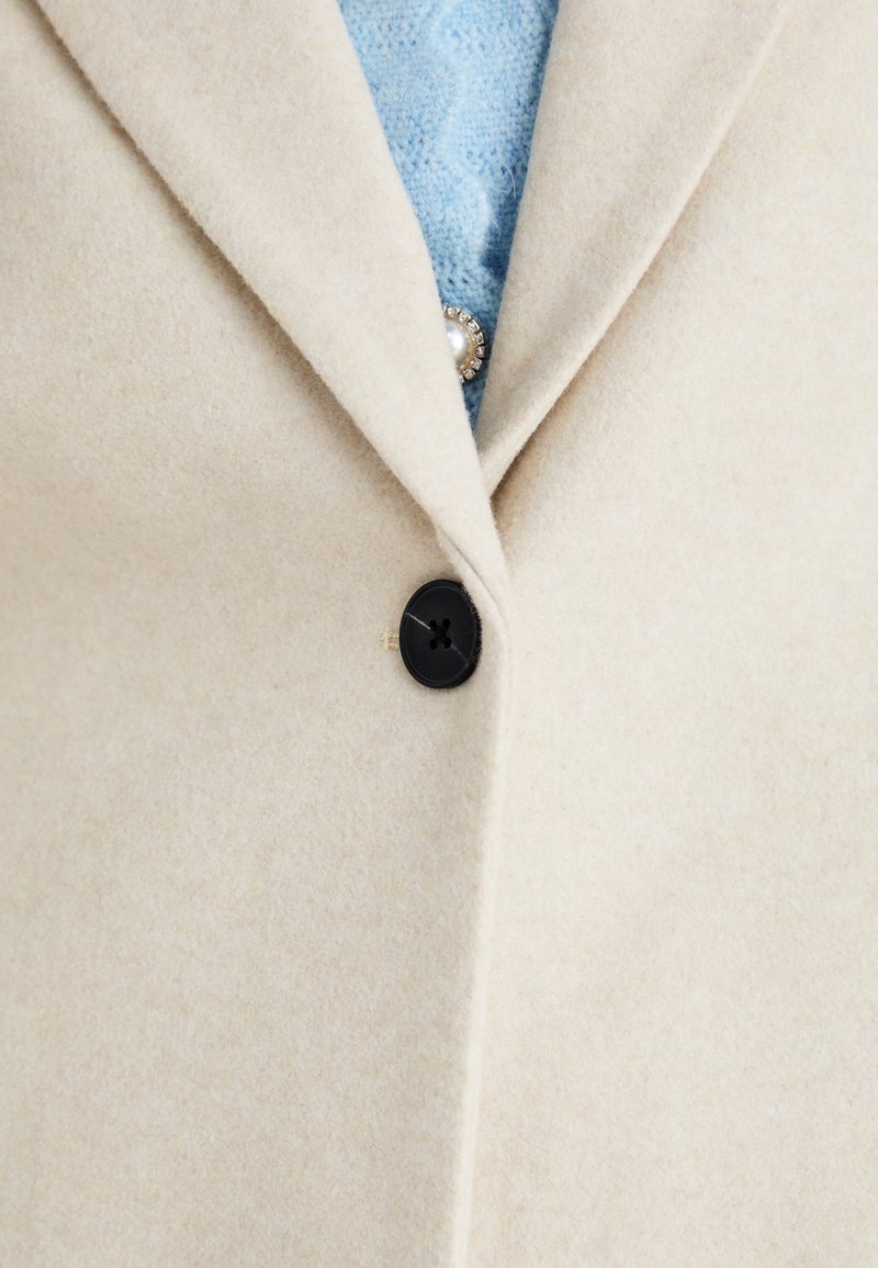 Beige Wollmantel mit schwarzem Knopfverschluss und großen Revers. Ein hellblaues Strickpullover ist darunter sichtbar, was den Kontrast der Texturen betont.