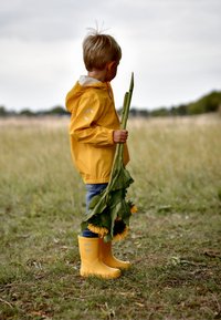 Gul vandtæt jakke, blå jeans og gule gummistøvler. Barn holder solsikker med grønne stilke og står på en græsmark.