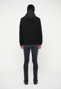 Crna textura pulover s kapuljačom, s rebrastim uzorcima; uparen s uskim tamnim trapericama i crnim cipelama, prikazano sa stražnje strane.