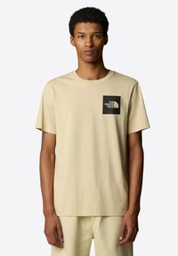 Beige bavlněné tričko s kulatým výstřihem, krátkými rukávy a černým čtvercovým logem na levém hrudníku s nápisem "THE NORTH FACE."
