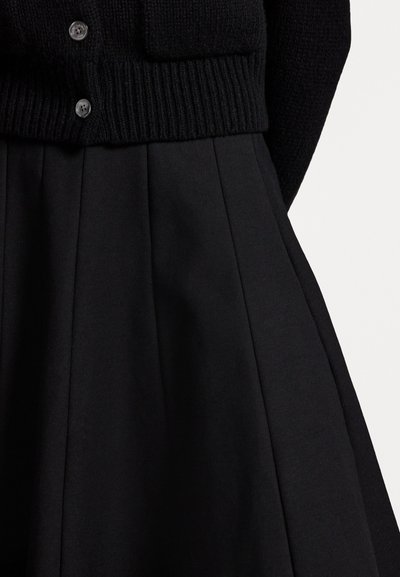 Čierny pletený cardigan s rebrovaným lemom, ktorý má päť gombíkov. Pod ním je plisovaná čierna sukňa s hladkým materiálom a štruktúrovaným dizajnom.