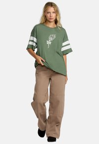 Groen oversized t-shirt met witte gestreepte mouwen en een bloemmotief. Gecombineerd met losse beige cargo broeken en zwarte schoenen.