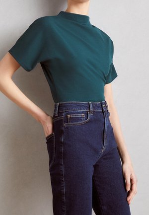 Persoon die een donkergroene top met korte mouwen draagt, ingestopt in een high-waisted donkerblauwe spijkerbroek, staand met één hand in de broekzak.