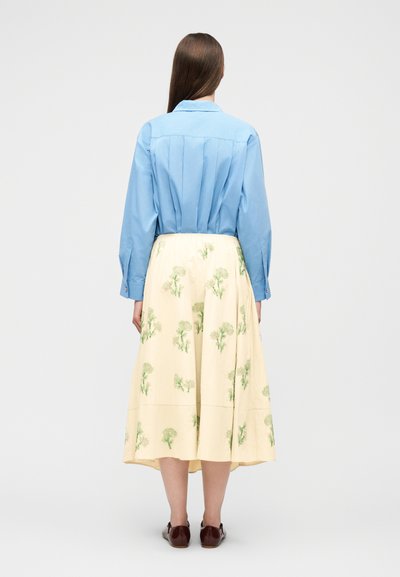 Γυναίκα με μακριά καστανά μαλλιά, που φορά μπλε πουκάμισο με κουμπιά και φούστα μίντι σε εκρού απόχρωση με πράσινα λουλουδάτα σχέδια, στέκεται με την πλάτη στραμμένη.