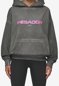 Sudadera gris de gran tamaño en tela suave con un bolsillo frontal. Presenta "PEGADOR" en letras brillantes y multicolores en el pecho.
