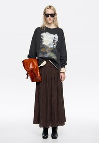 Sudadera negra de gran tamaño con un gráfico de montaña, blusa crema debajo, falda marrón de pliegues y bolso rojo brillante. Botas negras hasta el tobillo completan el look.