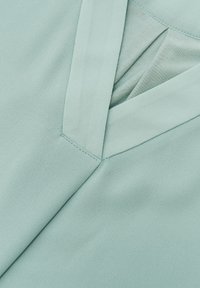 Blouse vert menthe clair avec un col en V, confectionnée dans un tissu doux et texturé. Elle présente des accents cousus et une silhouette fluide.