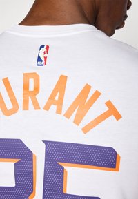 T-shirt bianco in cotone con la scritta "DURANT" in lettere maiuscole arancioni e "35" in viola con un motivo a pois, logo NBA nella zona del collo sul retro.