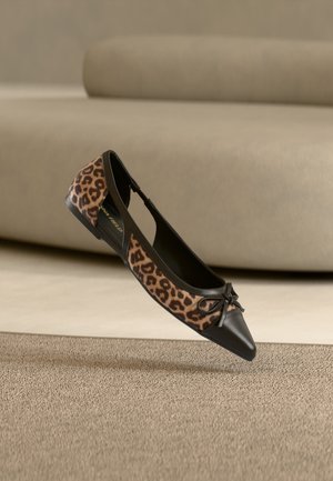 Chaussure plate avec côtés imprimé léopard, bout pointu noir et petit détail de nœud, présentée sur un fond de canapé beige et tapis.