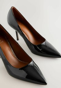 Fekete lakkbőr magassarkú cipők hegyes orral, vékony stiletto sarkú kialakítással és sima barna belső béléssel.