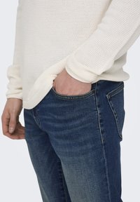 Weißer strukturierten Strickpullover mit langen Ärmeln, kombiniert mit blauen Jeans im klassischen Schnitt und leichten Ausbleichungen an den Oberschenkeln.