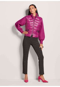 Eng gefütterte gesteppte Puffer-Weste in leuchtendem Lila, kombiniert mit einem lilafarbenen Strickpullover und schwarzen Hosen mit weißen Pünktchen. Helle pinke Pumps.