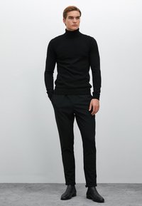 Zwarte coltrui met geribbelde textuur, gecombineerd met zwart tapse broek en zwarte enkellaarsjes, alles tegen een neutrale achtergrond.