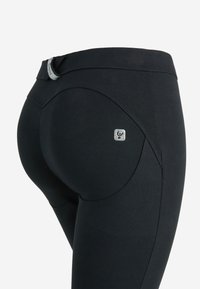 Mustat leggingsit, jotka on valmistettu joustavasta kankaasta, jossa on sileä pinta. Ne ovat kaarevamuotoiset ja sisältävät pienen logon vyötärön lähellä.