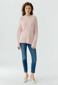 Frau trägt einen rosa gerippten Pullover, blaue Skinny-Jeans und beige spitze High Heels, steht vor einem schlichten hellen Hintergrund.