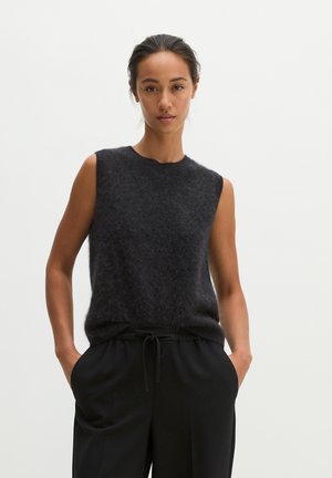 Jeune femme aux cheveux foncés attachés, portant un haut sans manches duveteux gris foncé et un pantalon noir, les mains dans les poches, debout devant un fond blanc.