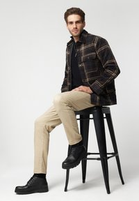 Kurtka w kratę w ciemnym brązie i beżu, czarna koszula, beżowe spodnie i czarne buty. Model siedzi na czarnym stołku na jasnym tle.