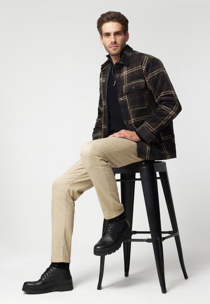 Kurtka w kratę w ciemnym brązie i beżu, czarna koszula, beżowe spodnie i czarne buty. Model siedzi na czarnym stołku na jasnym tle.