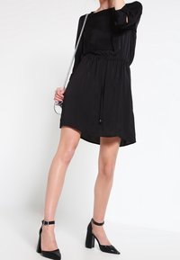 Vestido negro de manga larga con cintura elástica, largo hasta la rodilla y de tela ligera. Combinado con tacones negros de tira en el tobillo.