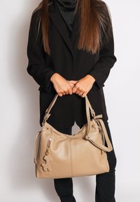 Beige leren handtas met twee handvatten, een ritssluiting, zijvakken en een speelse charm in de vorm van een beer eraan bevestigd.