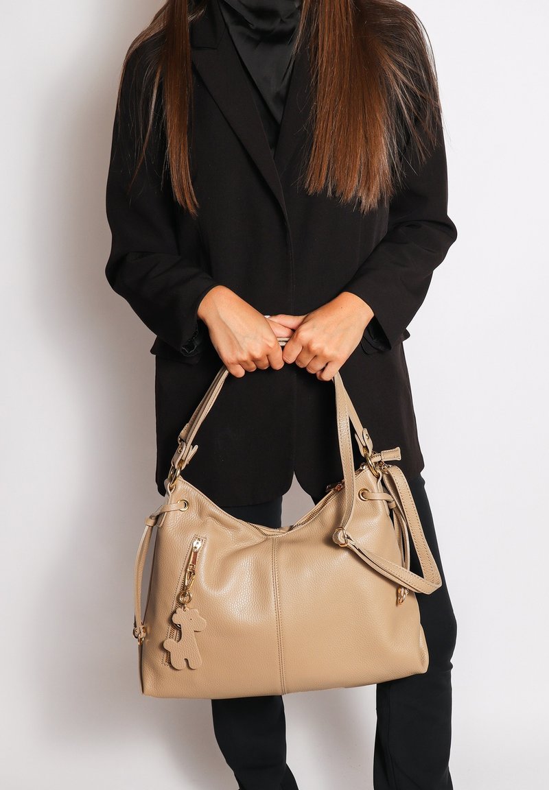 Beige leren handtas met twee handvatten, een ritssluiting, zijvakken en een speelse charm in de vorm van een beer eraan bevestigd.