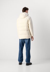Puffjacke in Off-White mit Kapuze, gestepptem Design und lässiger Passform. Getragen mit blauen Jeans und schwarzen Schuhen.