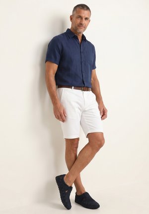 Mand lænende mod en væg iført navyblå kortærmet knap-skjorte, hvide shorts med brun bælte og navy slip-on sko.