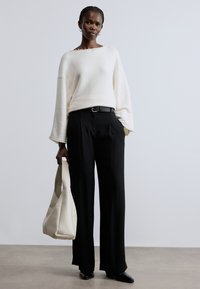 Fehér, túlméretezett pulóver harang alakú ujjakkal, magas derekú fekete nadrággal és krémszínű válltáskával párosítva. A megjelenést fekete cipő teszi teljessé.