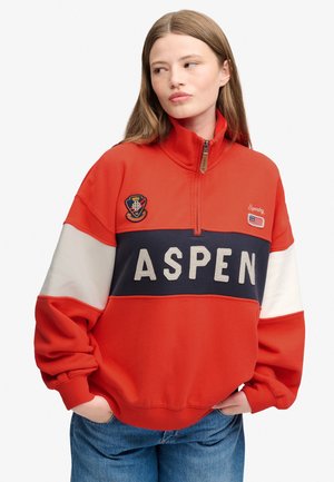 Ung kvinde iført en rød, hvid og marineblå pullover med teksten "ASPEN" og broderede patches, hænder i lommerne, kigger til siden.