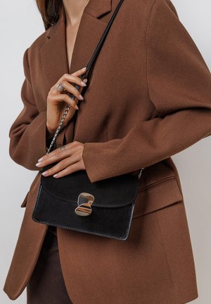 Czarna zamszowa torebka crossbody z zaokrągloną klapką i srebrnymi elementami, noszona na tle brązowego żakietu. Posiada srebrny łańcuszek jako pasek.