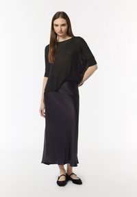 Czarna, przezroczysta dzianinowa bluzka z krótkim rękawem, w zestawie z długą, satynową spódnicą w ciemnym kolorze. Modelka ma na sobie czarne płaskie buty na pasek.