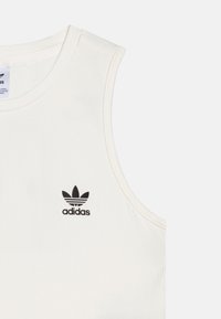 Λευκό αμάνικο μπλουζάκι φτιαγμένο από ριμπ ύφασμα με ένα μαύρο λογότυπο Adidas στο στήθος. Στρογγυλή λαιμόκοψη με ίσιες κοπές.