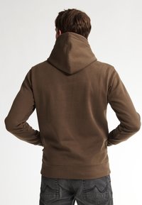 Bruine hoodie met een zachte textuur, voorzien van een opvallende capuchon en geribde manchetten, gecombineerd met donkere jeans. Eenvoudig, casual ontwerp.