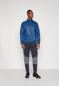 Navy lightweight jacket with a zip, paired with black and gray tapered pants featuring side accents, worn with black shoes.